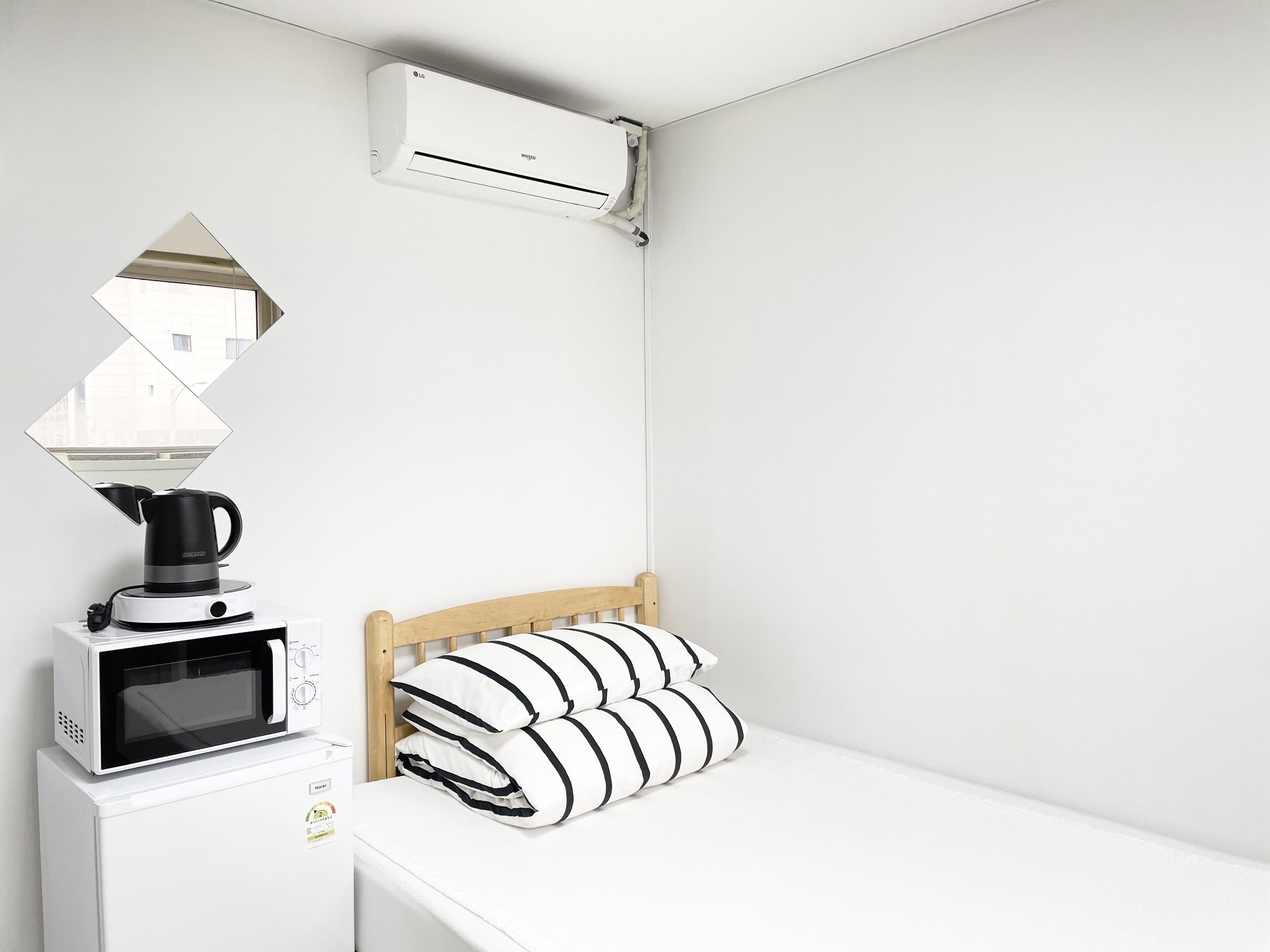 A compact room with a bed, a microwave, a kettle, and an air conditioner.