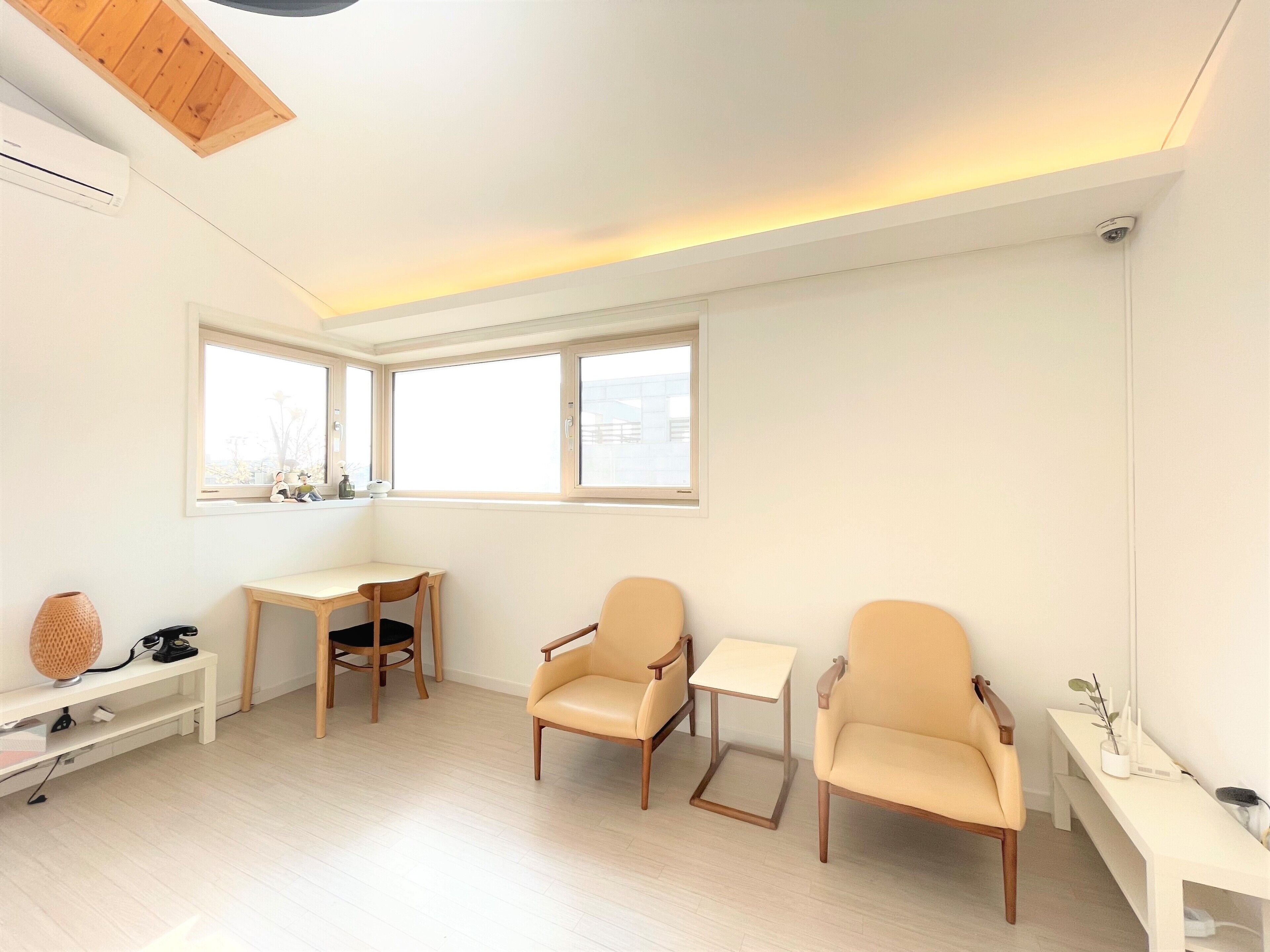 A modern, well-lit room with a wooden ceiling, a desk, two chairs, a small table, and a window with a view of the city.