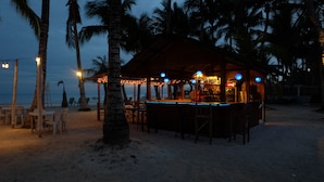 Un bar situé au bord de la plage, avec une structure en bois, entouré de palmiers et de chaises de plage.