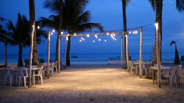 Une cérémonie de mariage organisée sur la plage, avec un auvent, des guirlandes lumineuses et des chaises en plastique blanches.