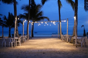 Decoração de casamento na praia, com toldo, luzes de Natal e cadeiras de plástico brancas.