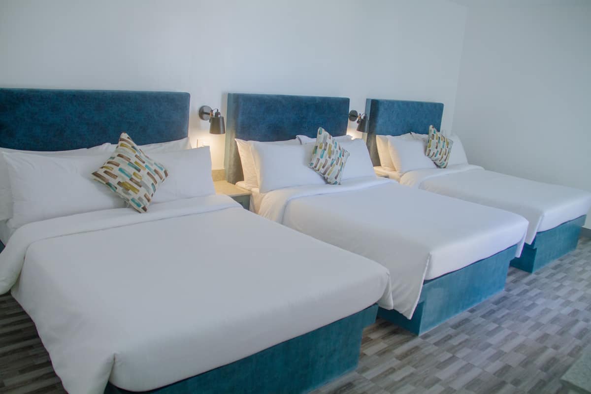 Quarto de hotel com três camas, cada uma com cabeceira azul e roupa de cama branca.