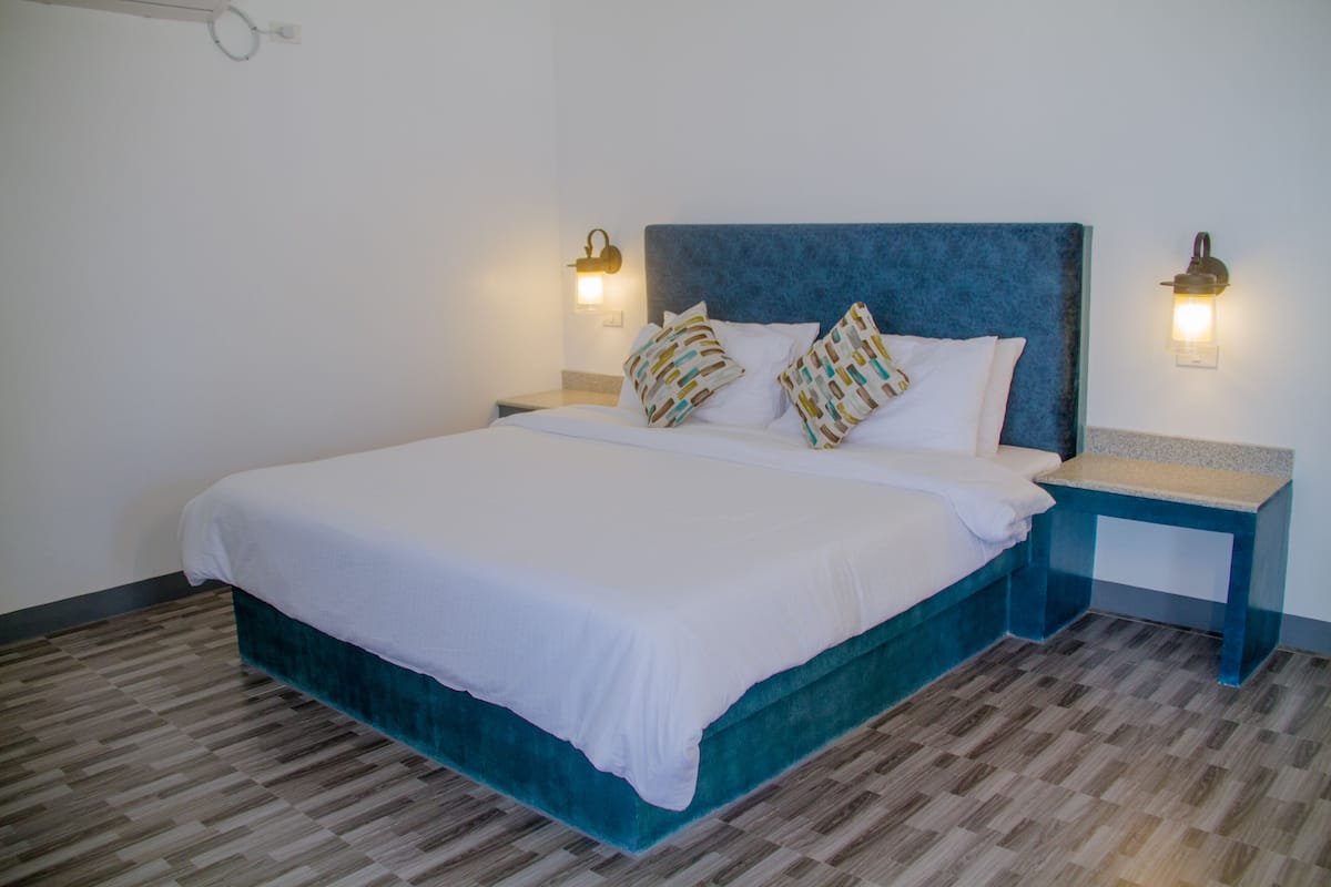 Cama bem arrumada com cabeceira verde-água, roupa de cama branca e almofadas estampadas. Duas mesas de cabeceira com luminárias fixadas na parede. O quarto tem piso de parquet de madeira.