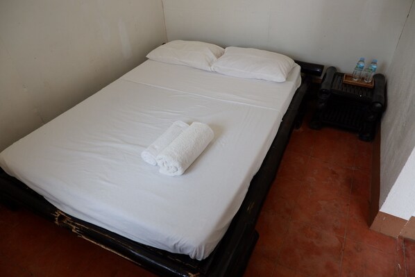 Uma cama de solteiro com roupa de cama branca e uma toalha enrolada, uma pequena mesa de cabeceira com garrafas de água e piso ladrilhado.