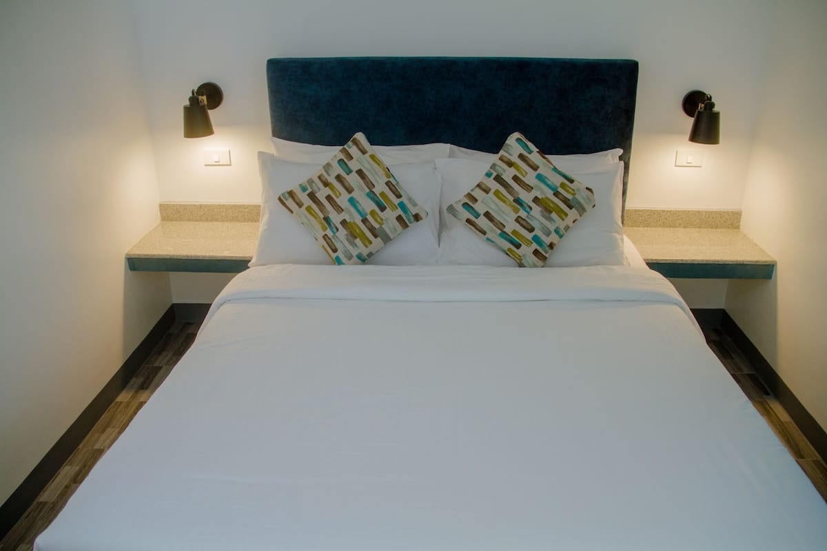 Uma cama bem arrumada com cabeceira azul, duas mesas de cabeceira com luminárias e almofadas estampadas.