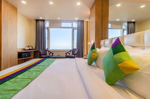 Quarto de hotel com uma cama grande, uma escrivaninha com cadeira, vista para o exterior por uma janela e uma mesa de cabeceira com abajur.