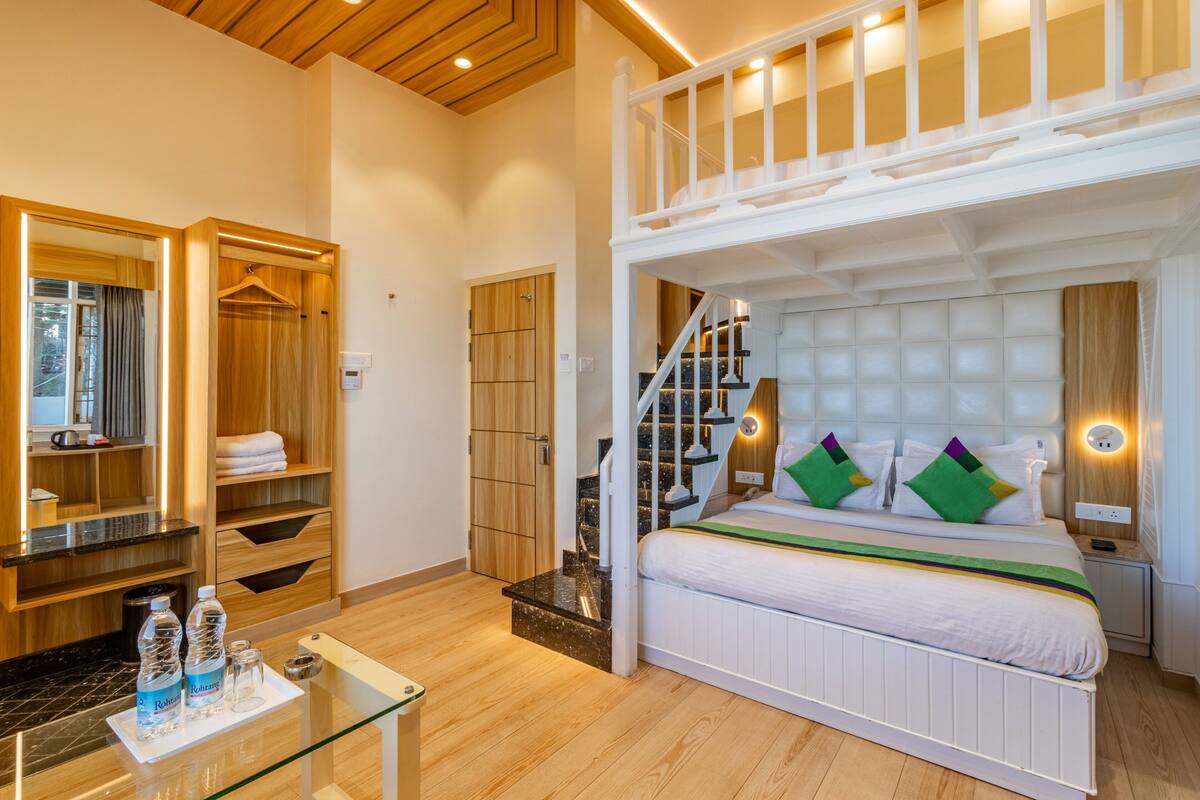 A hotel room with a bunk bed, a wooden wardrobe, a glass table with toiletries, and a staircase leading to an upper level.