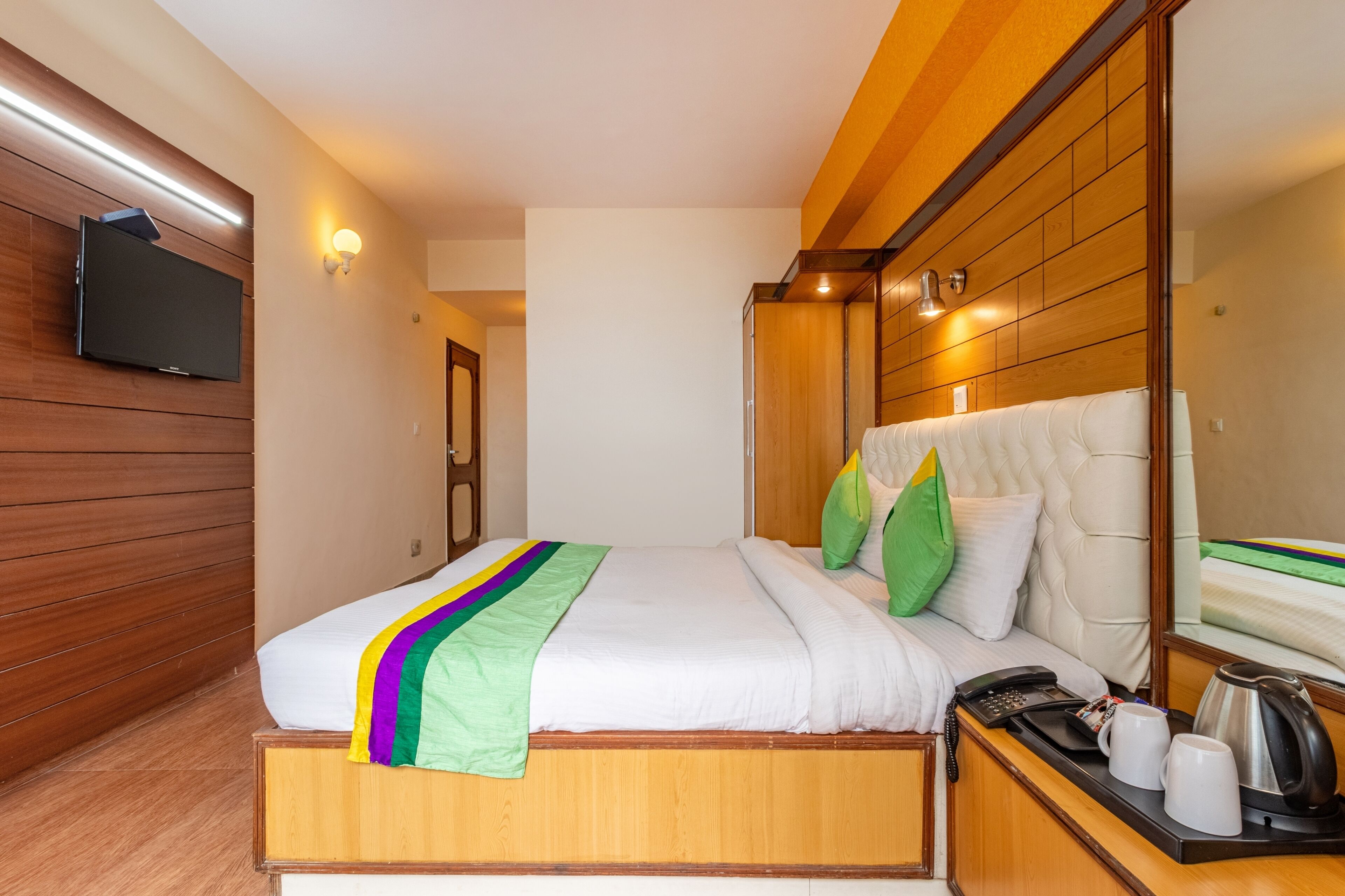 A hotel room with a wooden bed, a flat-screen TV mounted on the wall, a bedside table with a phone, and a mirror.