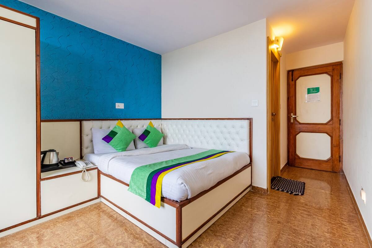 A bedroom with a white bed, green and purple pillows, a blue accent wall, a wooden door, and a small bedside table with a kettle and phone.