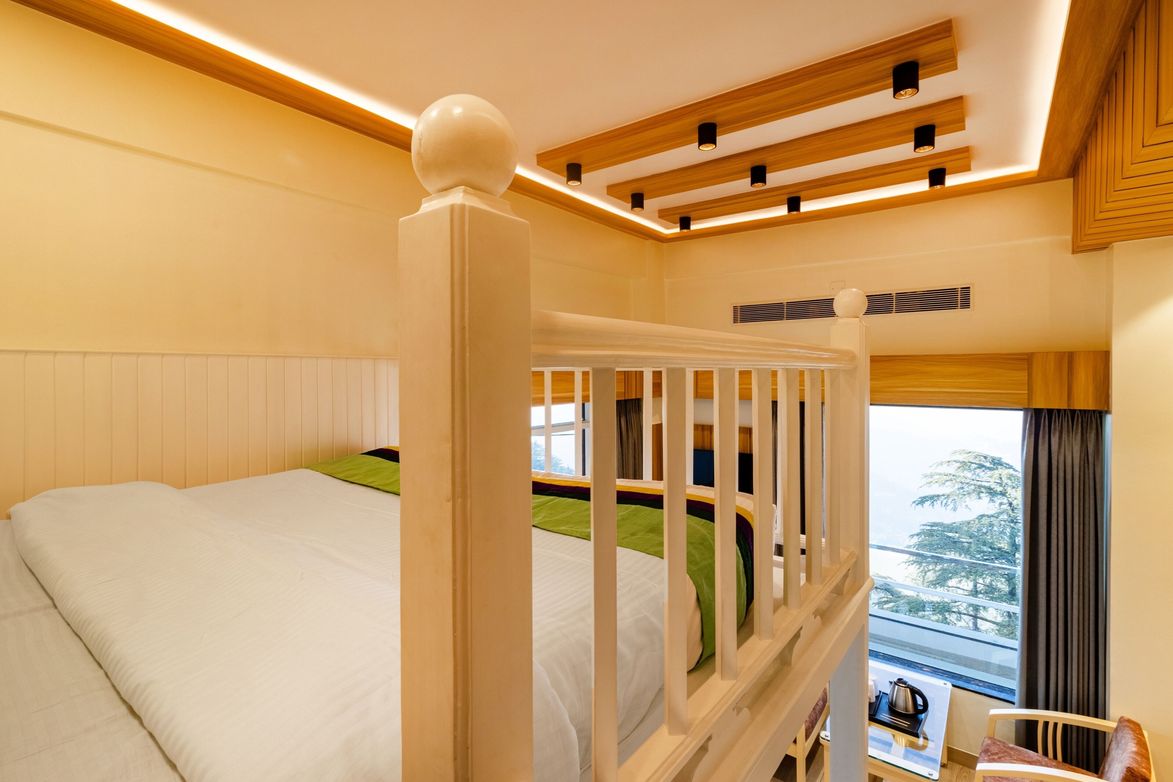 A hotel room with a wooden bed frame, a view of the outdoors, and a small table with a kettle.