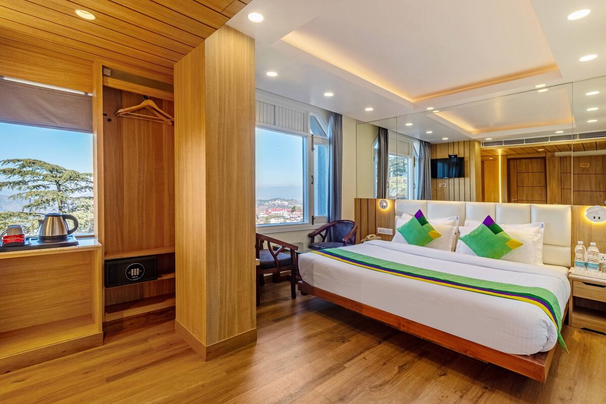 A modern hotel room with a wooden floor, a large bed with colorful pillows, a desk with a kettle, and a wardrobe.