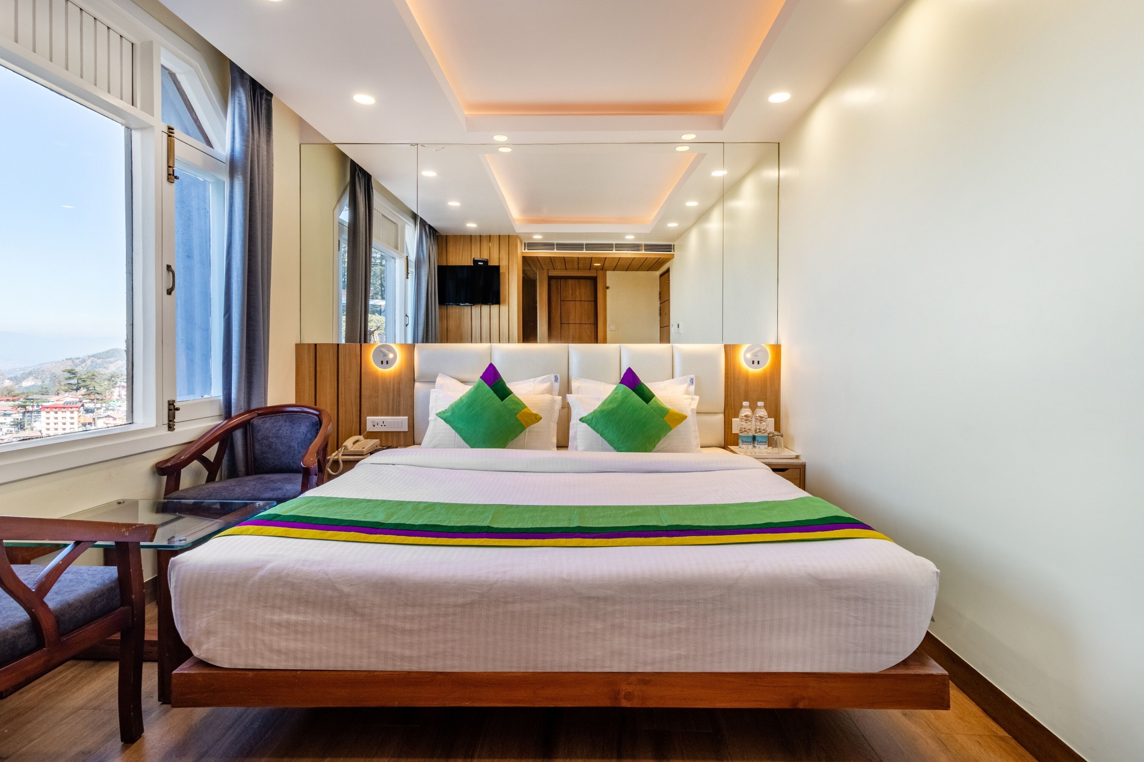 A modern hotel room with a large bed, a desk, and a chair. There is a window with curtains, a flat-screen TV, and a mirror on the wall.