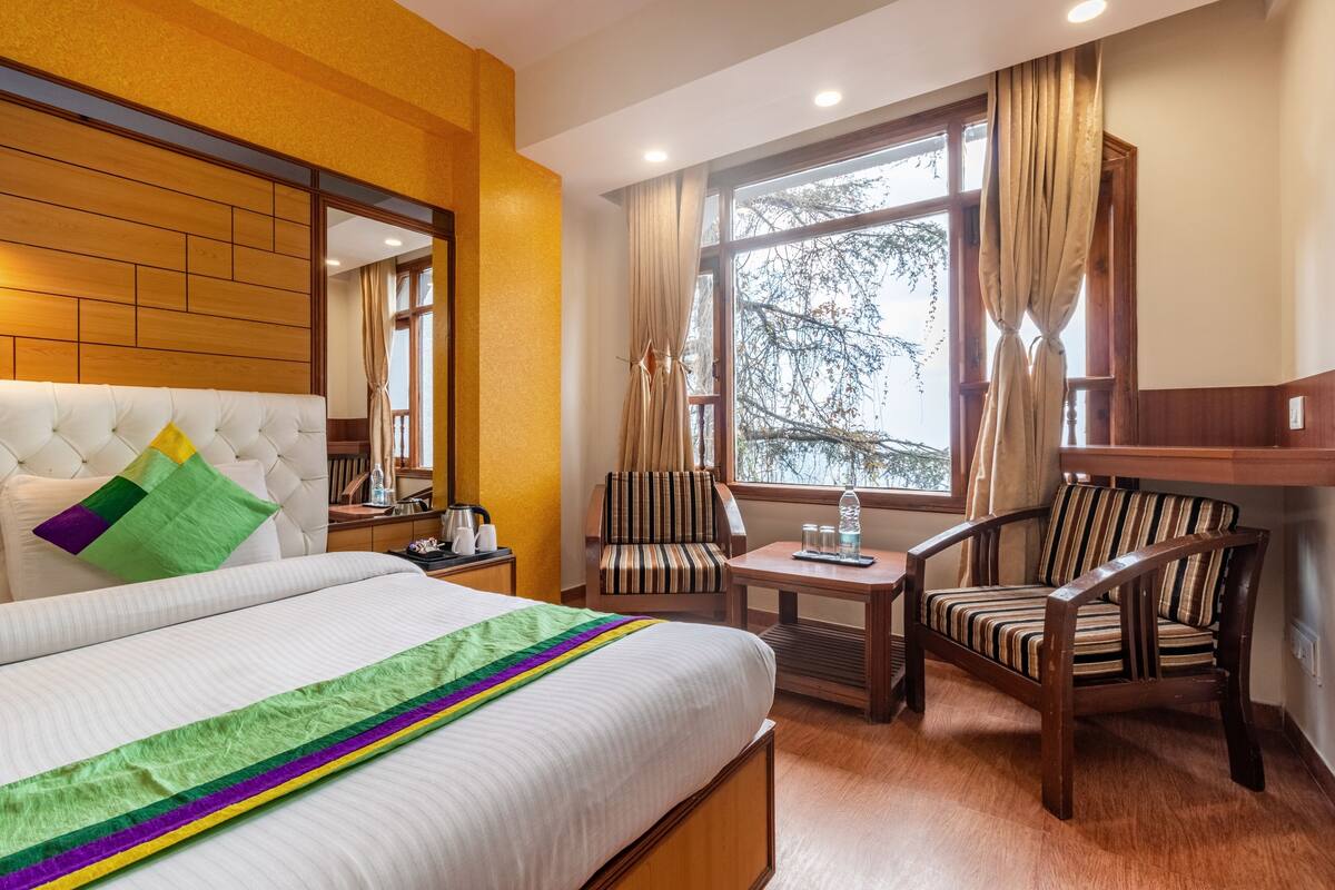 A hotel room with a large bed, a wooden headboard, a striped armchair, a wooden side table, and a window with curtains.