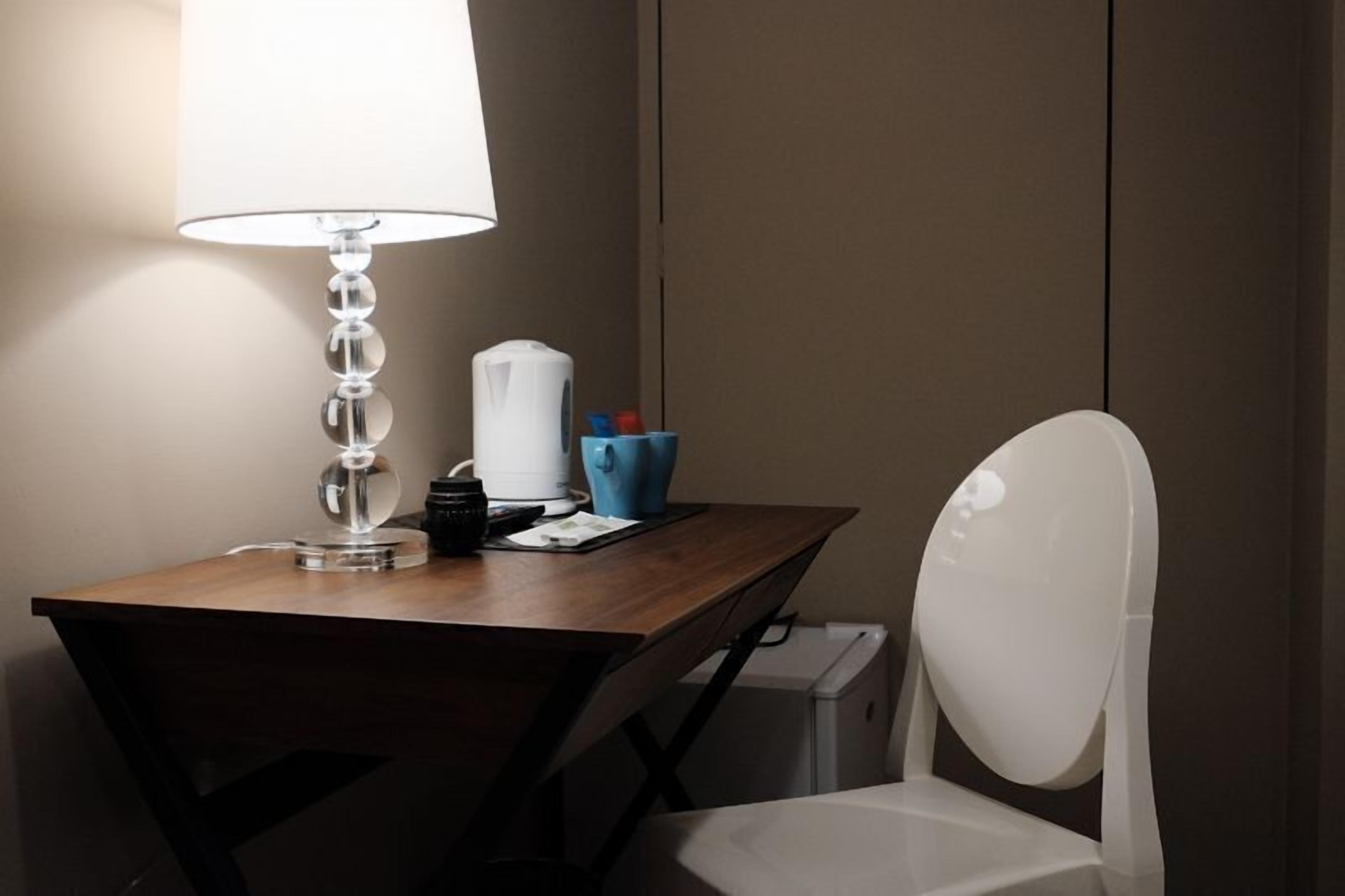 Une chambre d’hôtel avec un bureau en bois, une chaise blanche, une lampe, une machine à café et un petit récipient bleu.