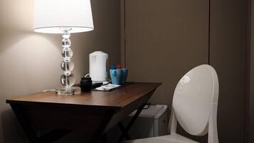 Une chambre d’hôtel avec un bureau en bois, une chaise blanche, une lampe, une machine à café et un petit récipient bleu.
