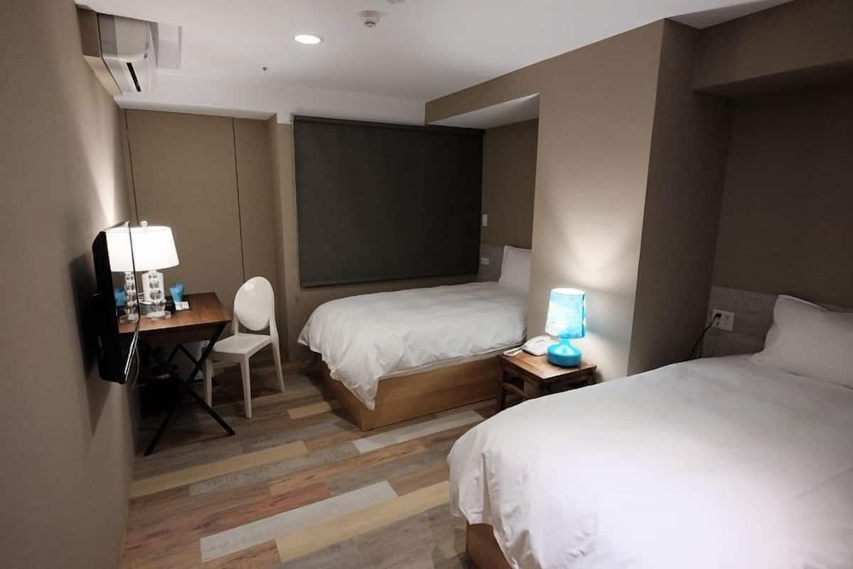 A hotel room with two beds, a desk with a chair, a TV, and a window with a blind.