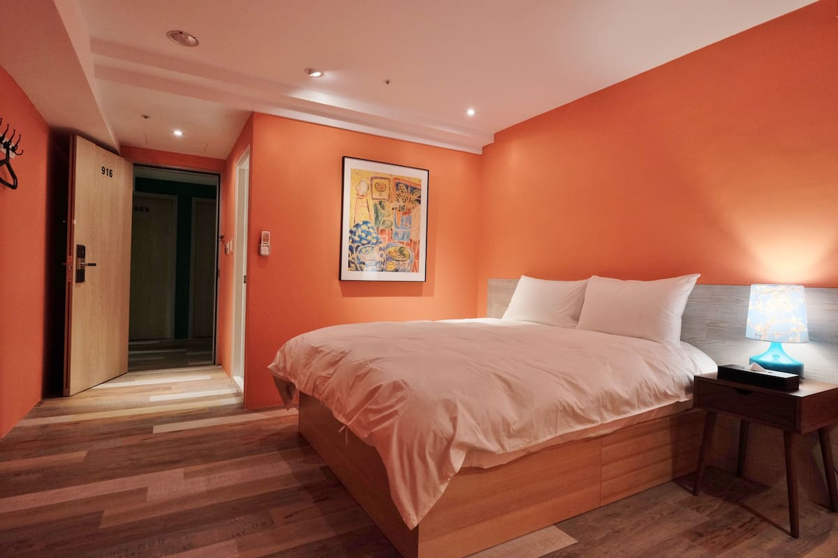 A bedroom with a large bed, a nightstand with a lamp, and a door leading to another room.
