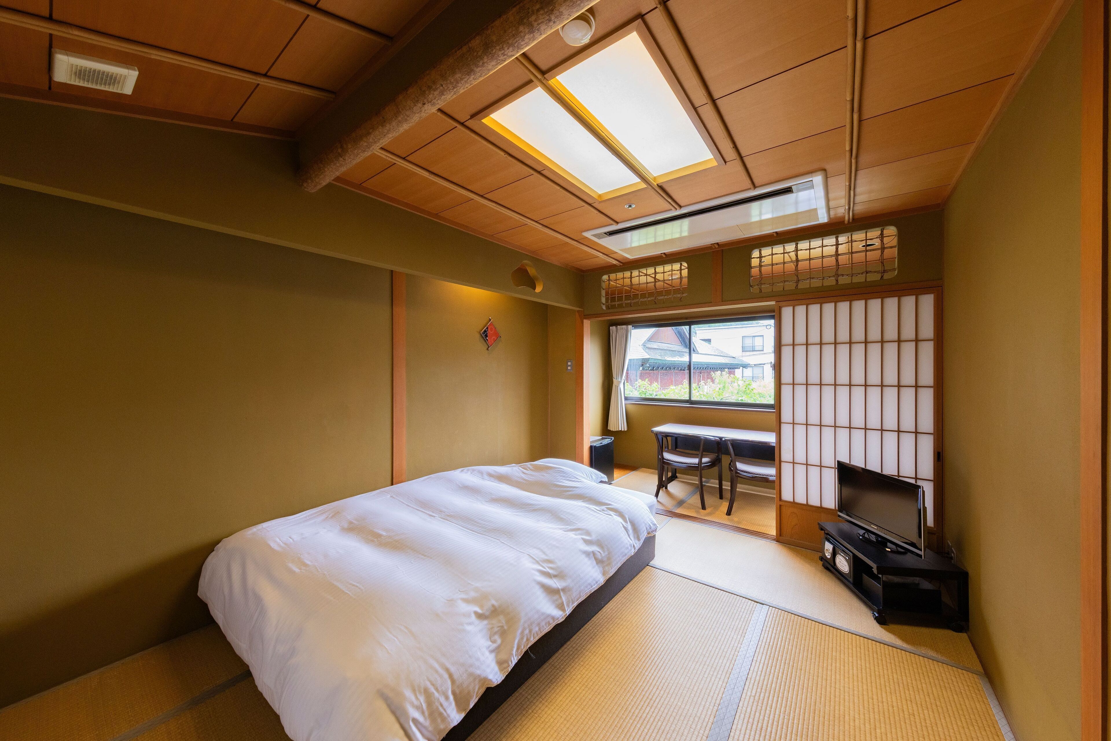 Una habitación tradicional japonesa con suelo de tatami, una cama, un televisor sobre un soporte y una puerta corredera con vistas al exterior.