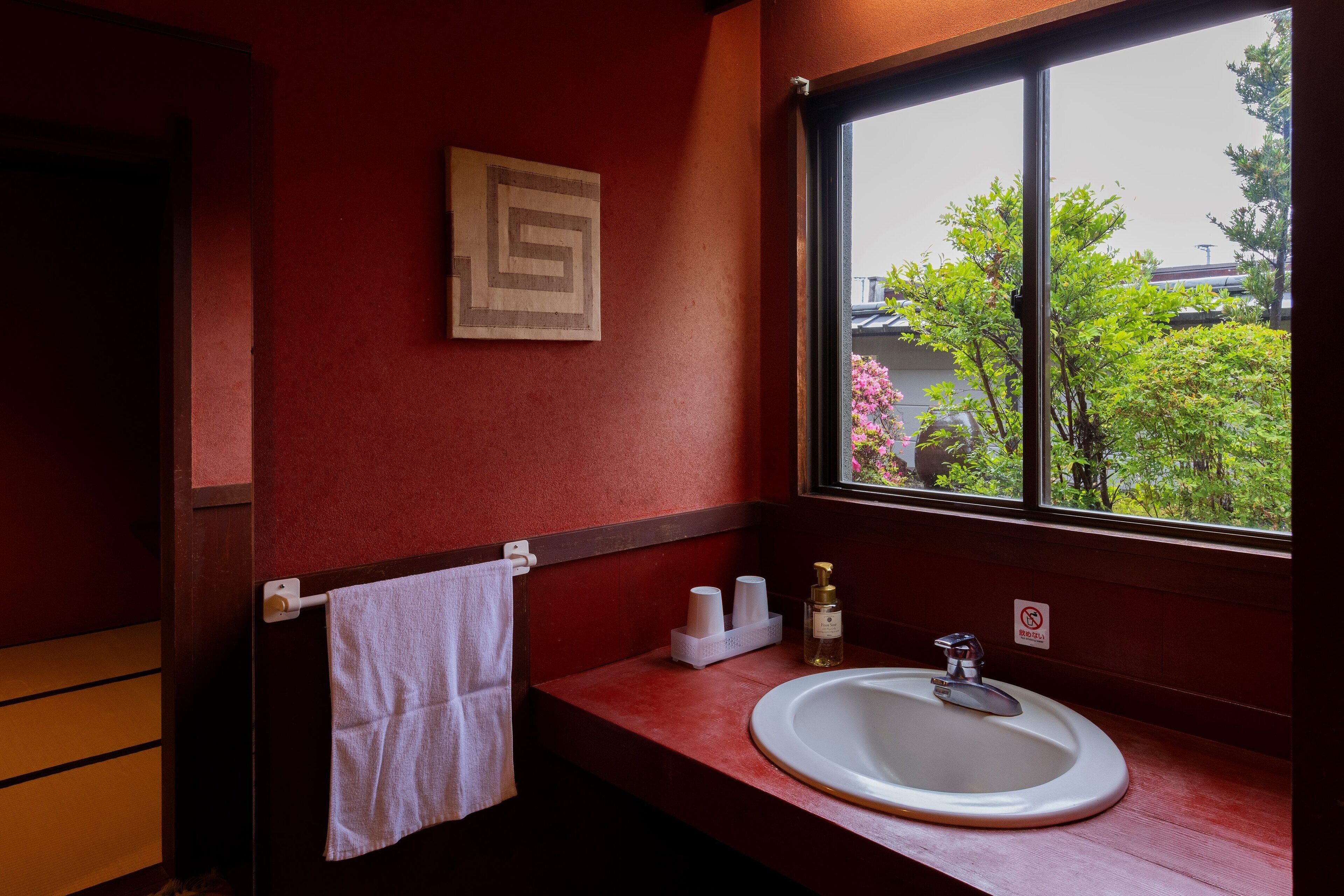Ein Badezimmer mit roter Wand, weißem Waschbecken und einem Fenster mit Blick auf Grünflächen und Gebäude.