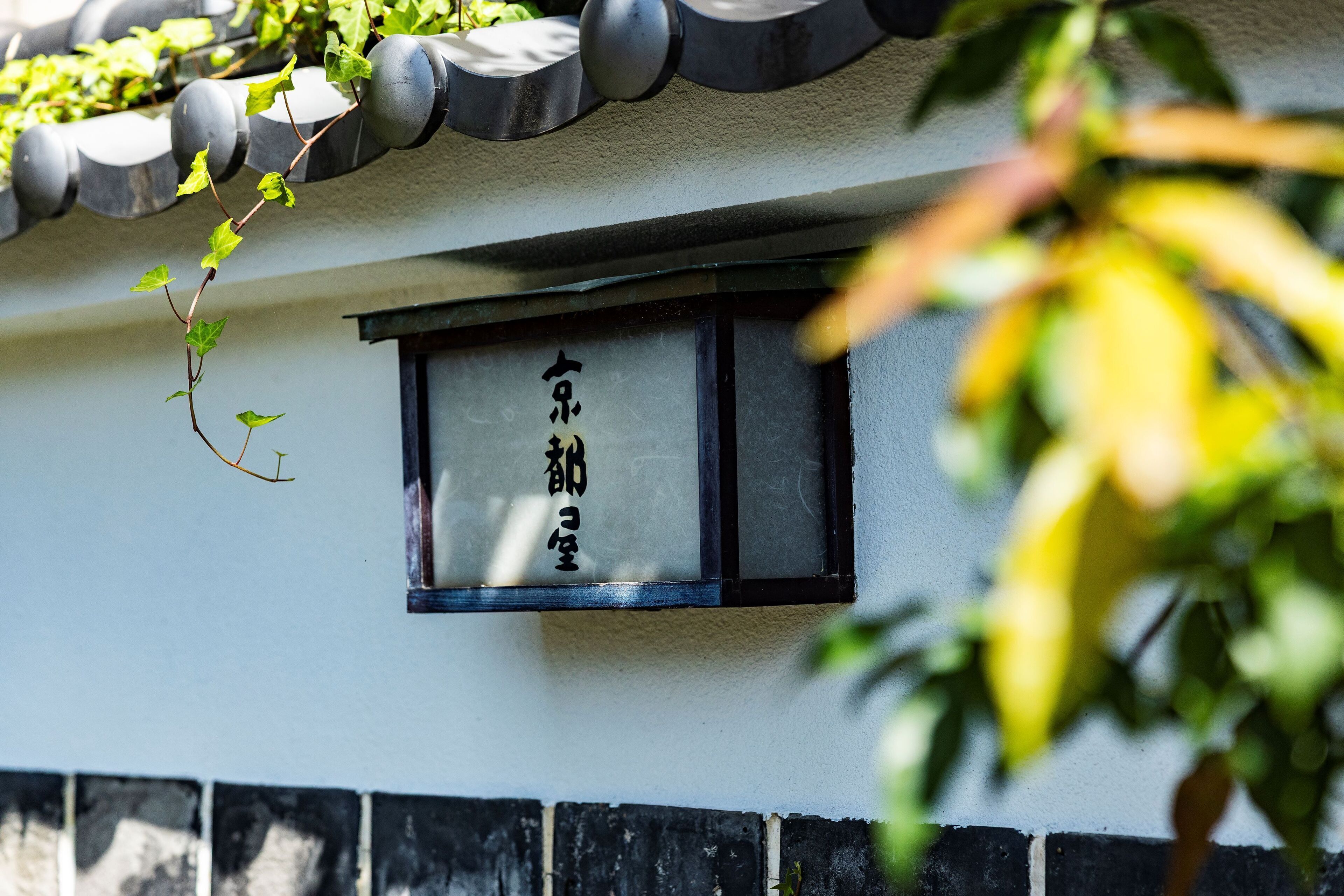 Un bâtiment traditionnel avec un toit recouvert de tuiles, une fenêtre aux montants noirs et une enseigne ornée de caractères japonais.