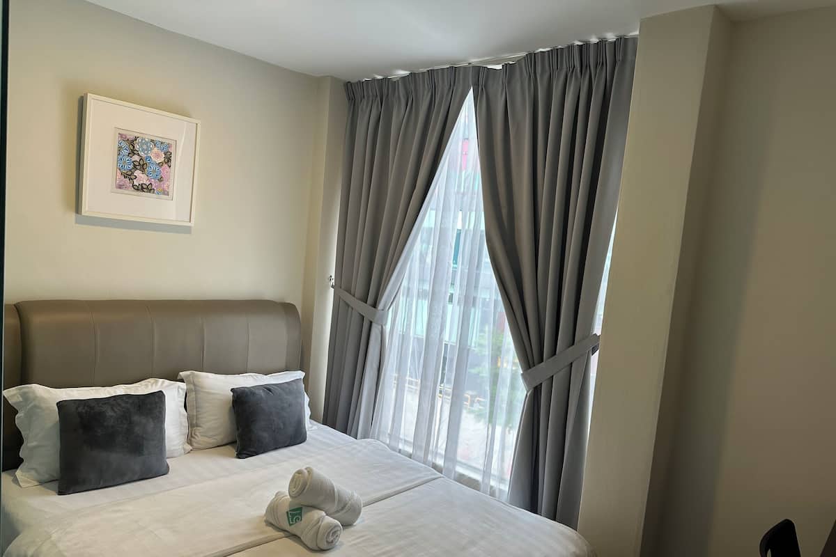 Quarto de hotel com cama, almofadas, cabeceira, janela com cortinas e um quadro na parede.