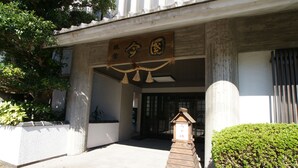 Entrada de um edifício com uma placa de madeira, uma pequena estrutura de madeira e uma sebe bem cuidada.