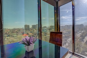 Habitación con un ventanal amplio que ofrece vistas a la ciudad, una silla de madera y una planta en maceta sobre la mesa.