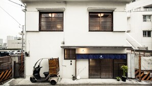Un edificio de dos pisos con fachada blanca, una pequeña entrada a tienda con toldo azul y una moto estacionada.