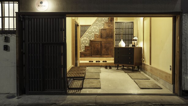 Una entrada tradicional japonesa con un portón de madera, una escalera que conduce a una habitación con un armario de madera y un jarrón.