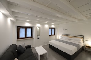 A bedroom with a bed, a small table, a couch, and two windows with curtains. - Piano Tre (Verona)