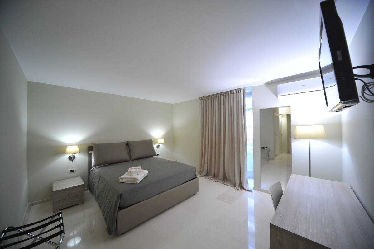 A modern hotel room with a bed, bedside tables, a desk, and a flat-screen TV mounted on the wall.