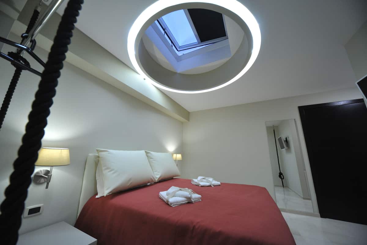 A hotel room with a bed, a circular skylight, a bedside table with a lamp, and a door.