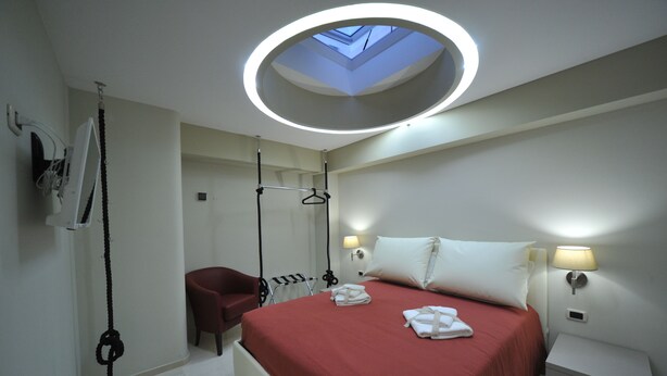 A modern hotel room with a skylight, a bed with red bedding, a red armchair, a small desk with a lamp, and a coat rack.