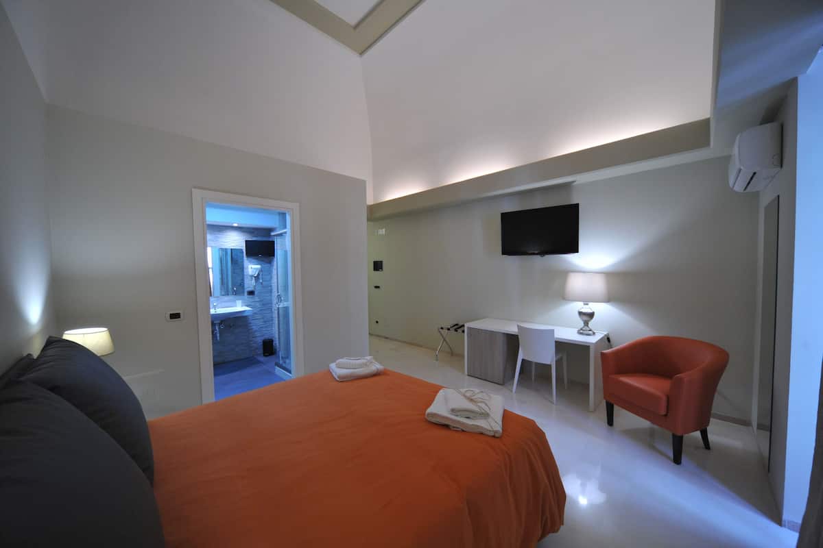 A hotel room with a bed, a desk, a chair, a TV, and a bathroom visible through an open door.