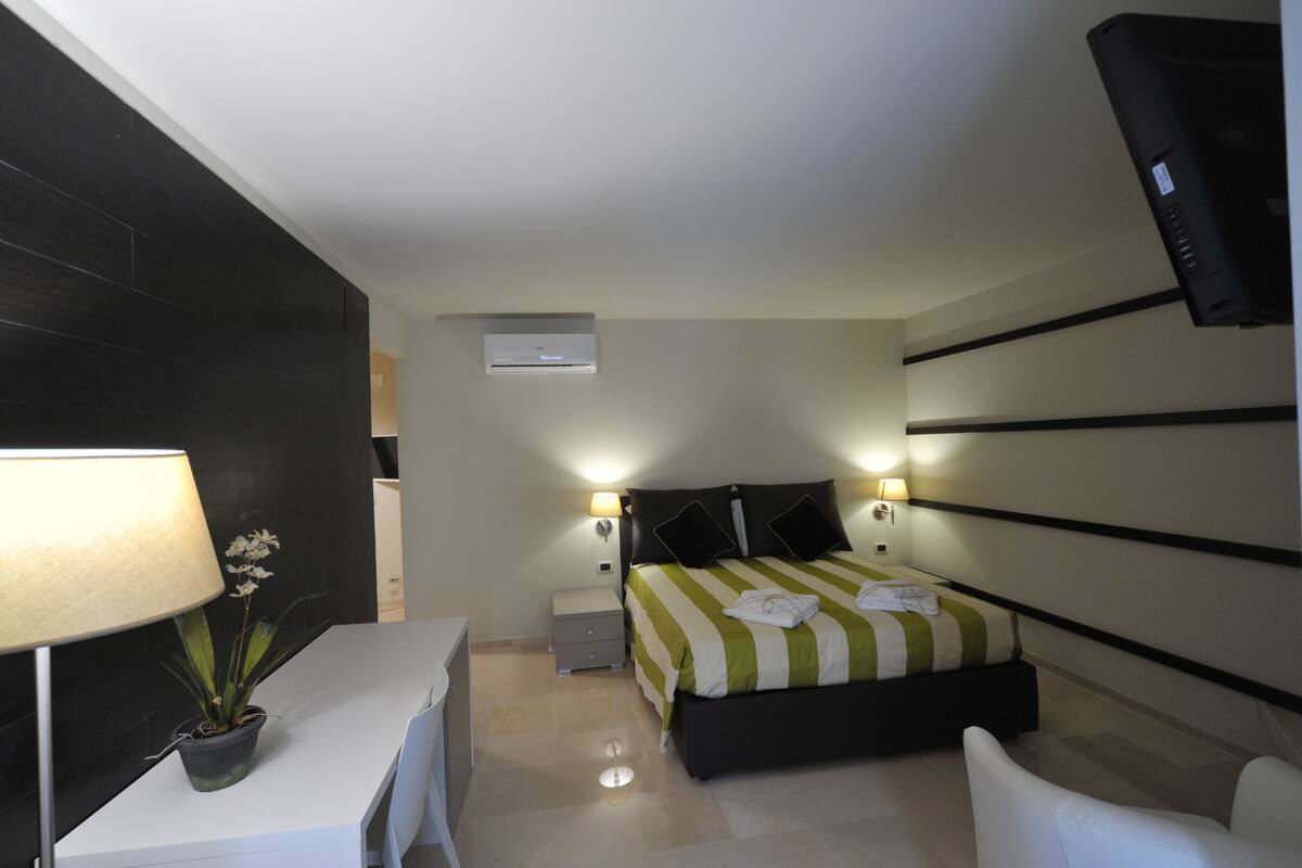 A modern hotel room with a bed, bedside tables, a desk, a chair, a TV, and a potted plant.
