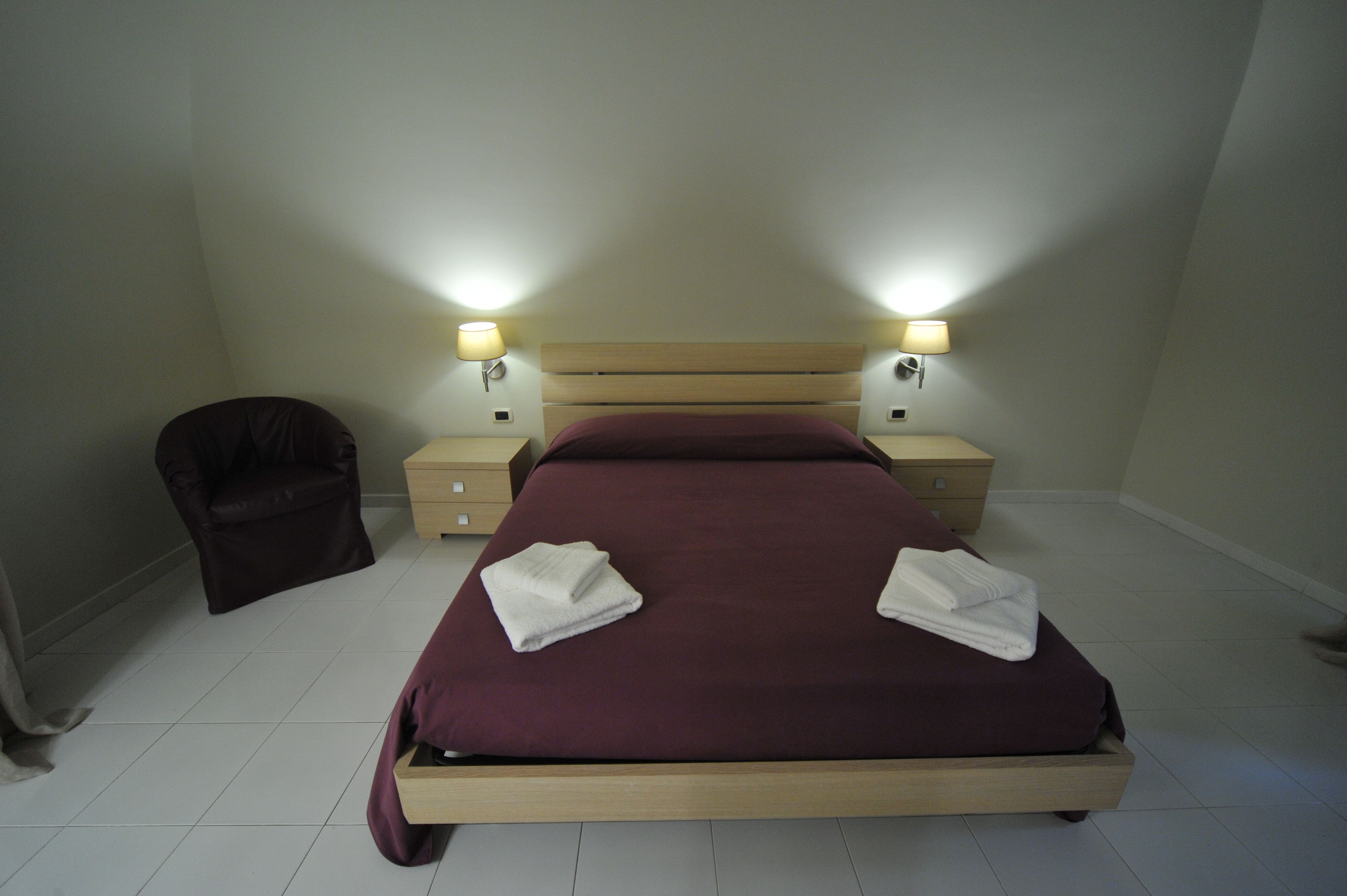 Uma cama bem arrumada com estrutura de madeira, duas luminárias de cabeceira, uma cadeira e uma mesinha de apoio.
