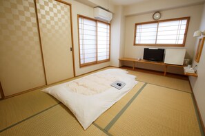 Una habitación con suelo de tatami, una cama con edredón blanco, un televisor sobre un soporte de madera y una mesita con un teléfono y un control remoto.