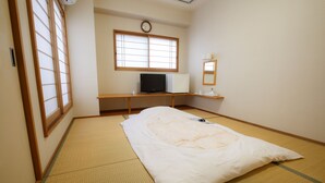 Une pièce avec un sol recouvert de tatamis, un lit avec un matelas et un oreiller blancs, un bureau avec un ordinateur, un petit réfrigérateur, une horloge et un miroir au mur.