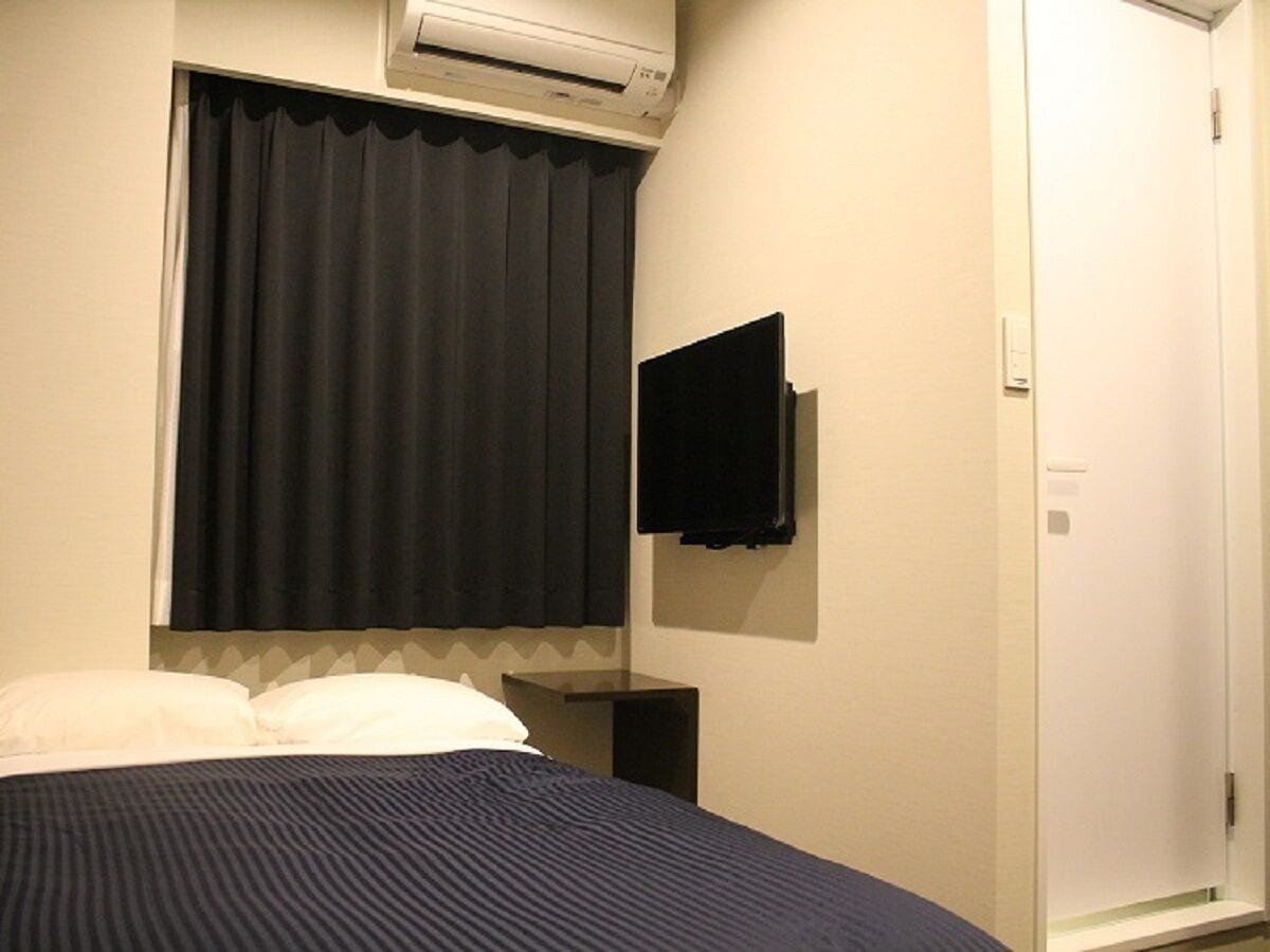 A hotel room with a bed, a flat-screen TV mounted on the wall, a window with curtains, and an air conditioning unit above the window.