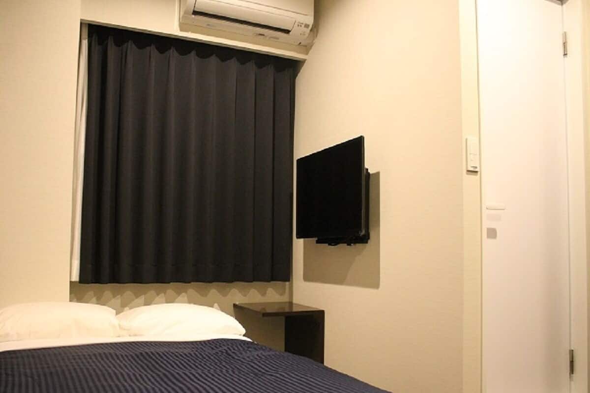 Habitación de hotel con cama, televisor de pantalla plana en la pared, ventana con cortinas y unidad de aire acondicionado sobre la ventana.