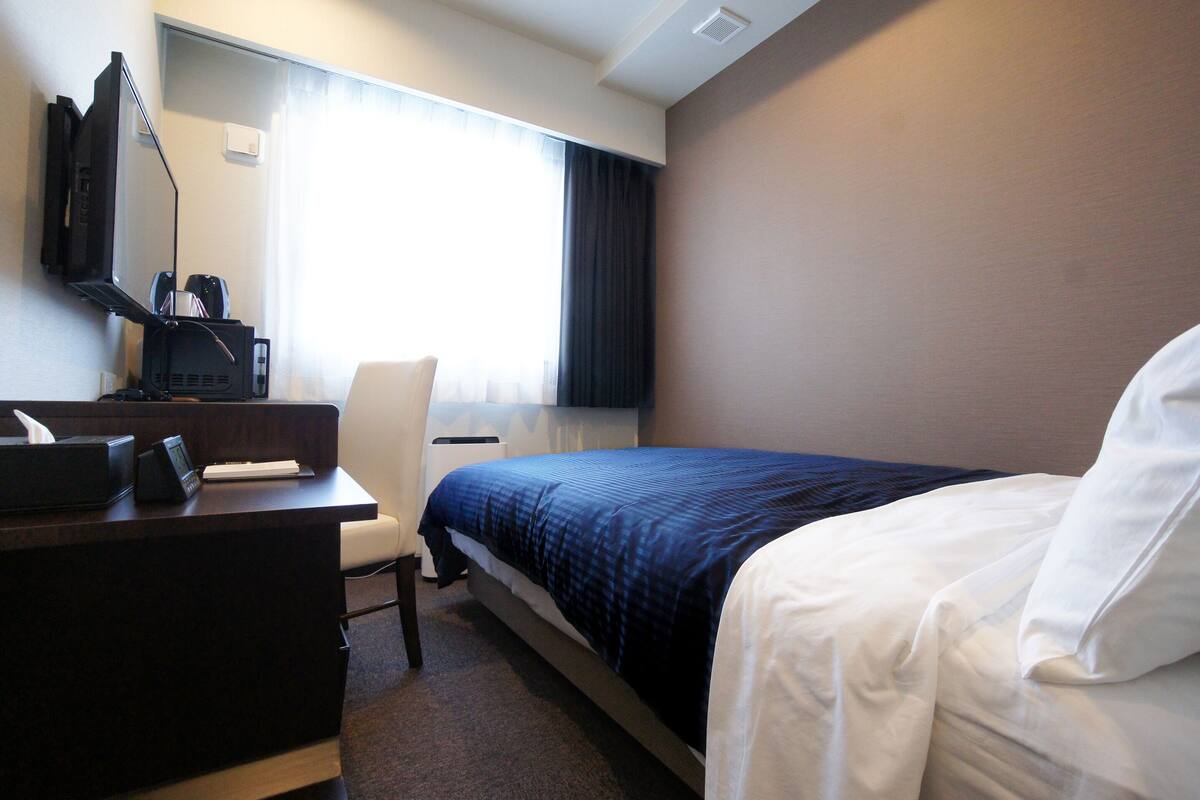 Habitación de hotel con cama, escritorio, silla, televisor y ventana con cortinas.