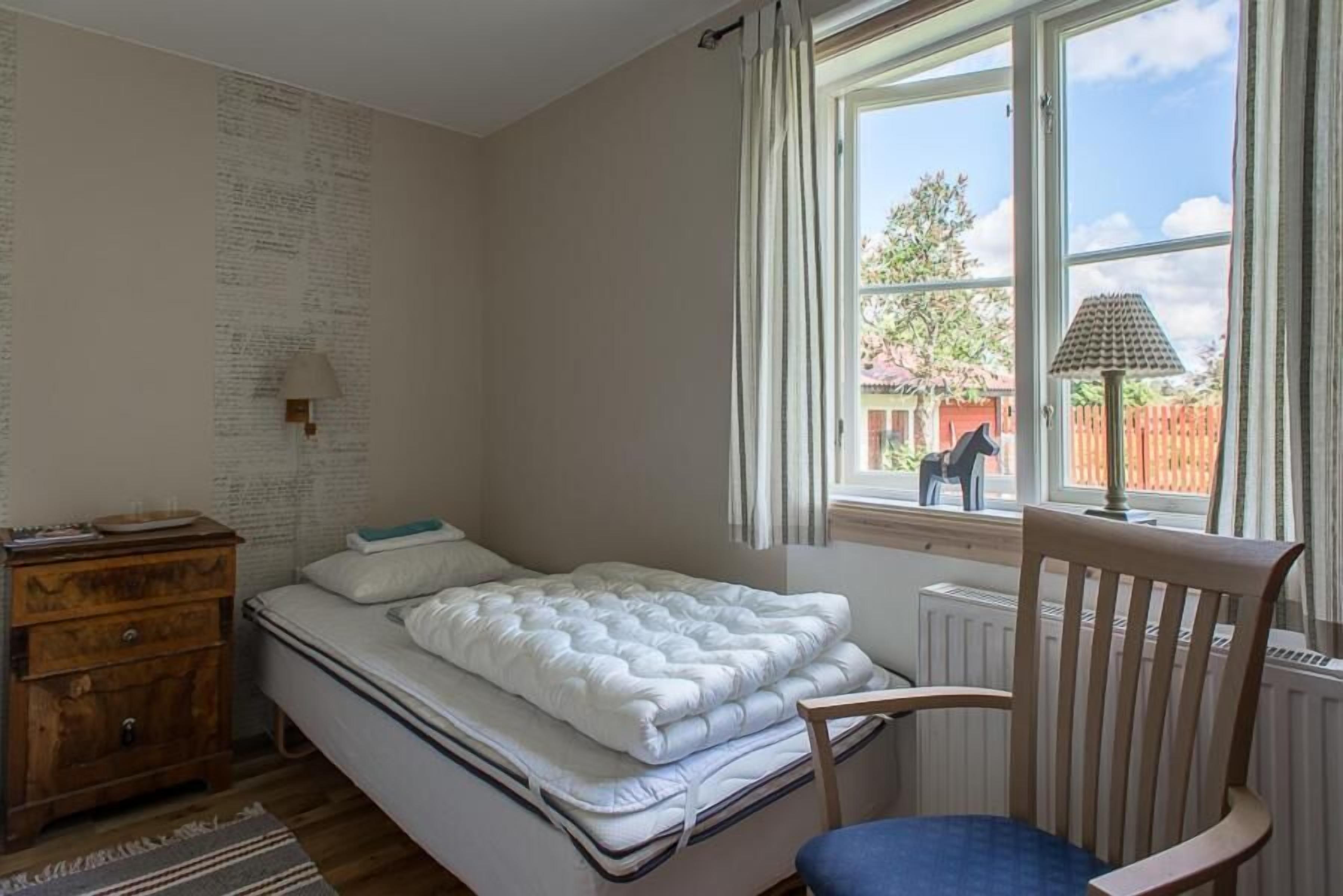 A bedroom with a bed, a wooden chair, a nightstand, and a window with curtains.