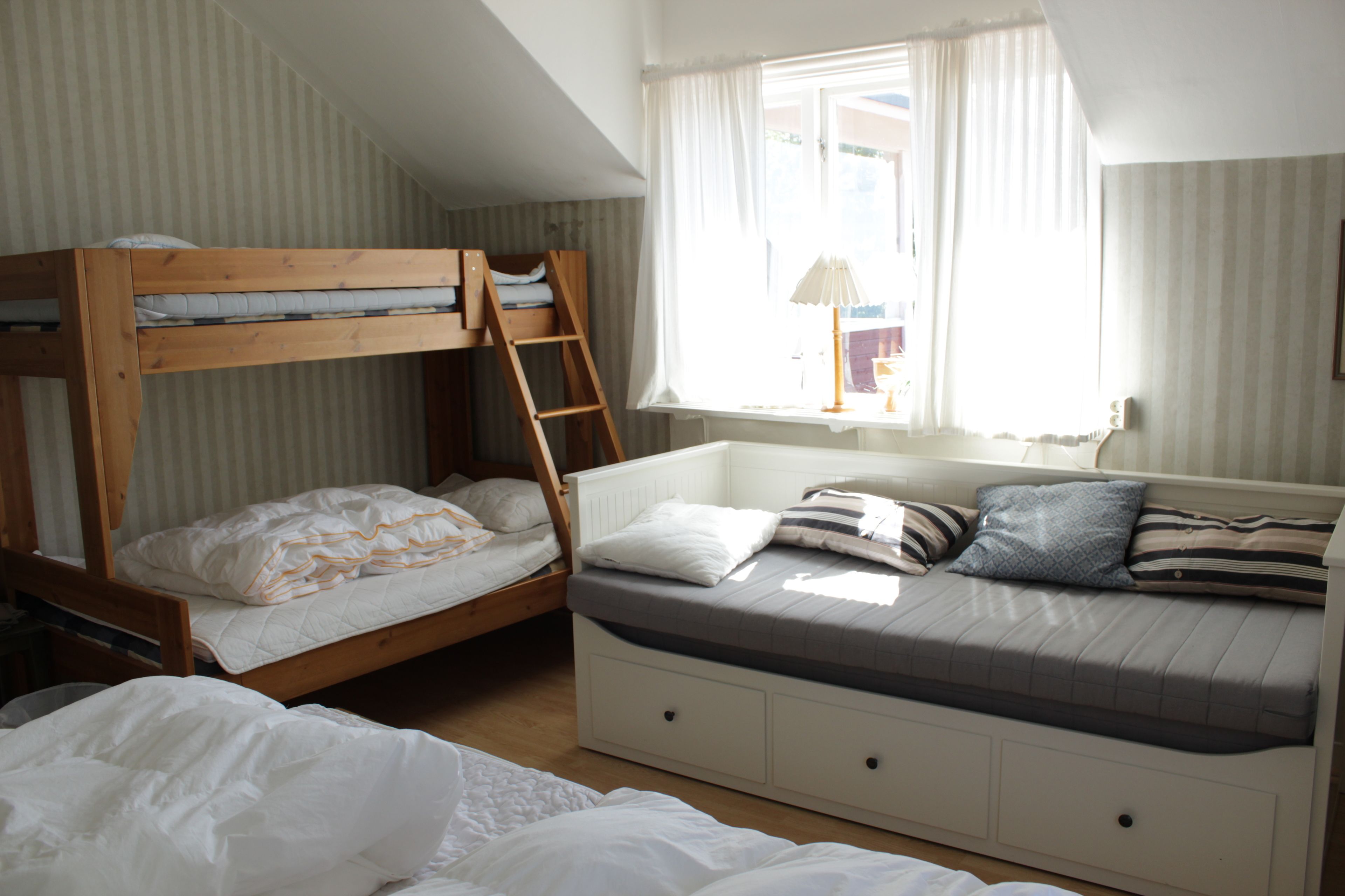 A dorm-style room with bunk beds and a daybed, featuring a window with curtains and a lamp on the nightstand.