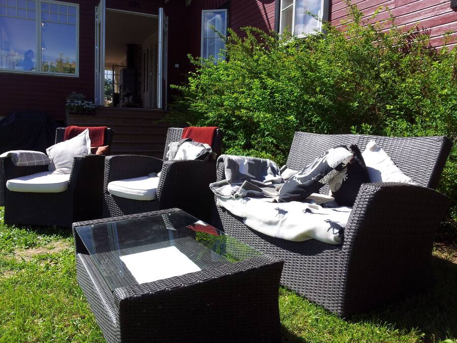 An outdoor seating area with wicker furniture and a glass table, surrounded by greenery and a red building in the background.