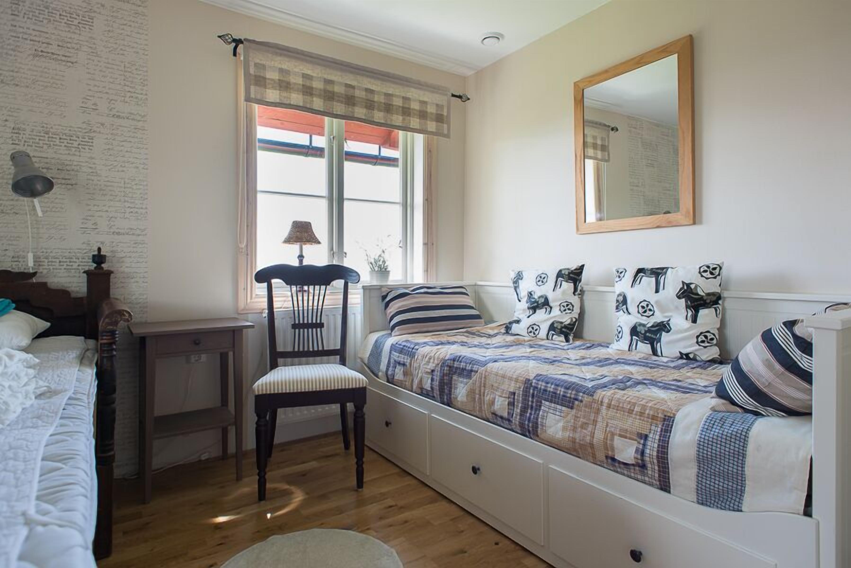 Un dormitorio con cama, una silla, un escritorio, una ventana con cortinas y un espejo en la pared.
