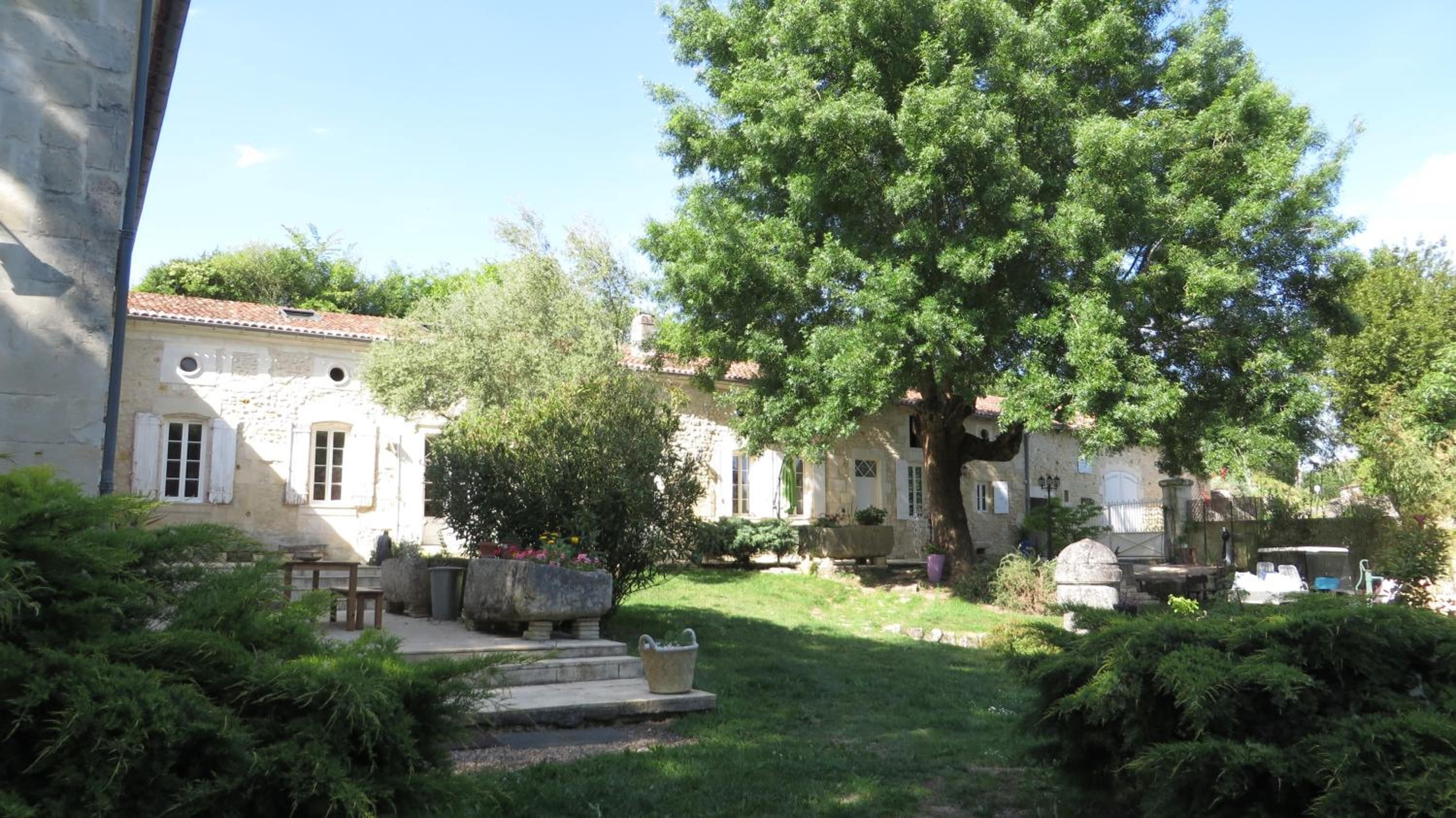 Un bâtiment en pierre avec un jardin, un grand arbre et un banc en pierre.