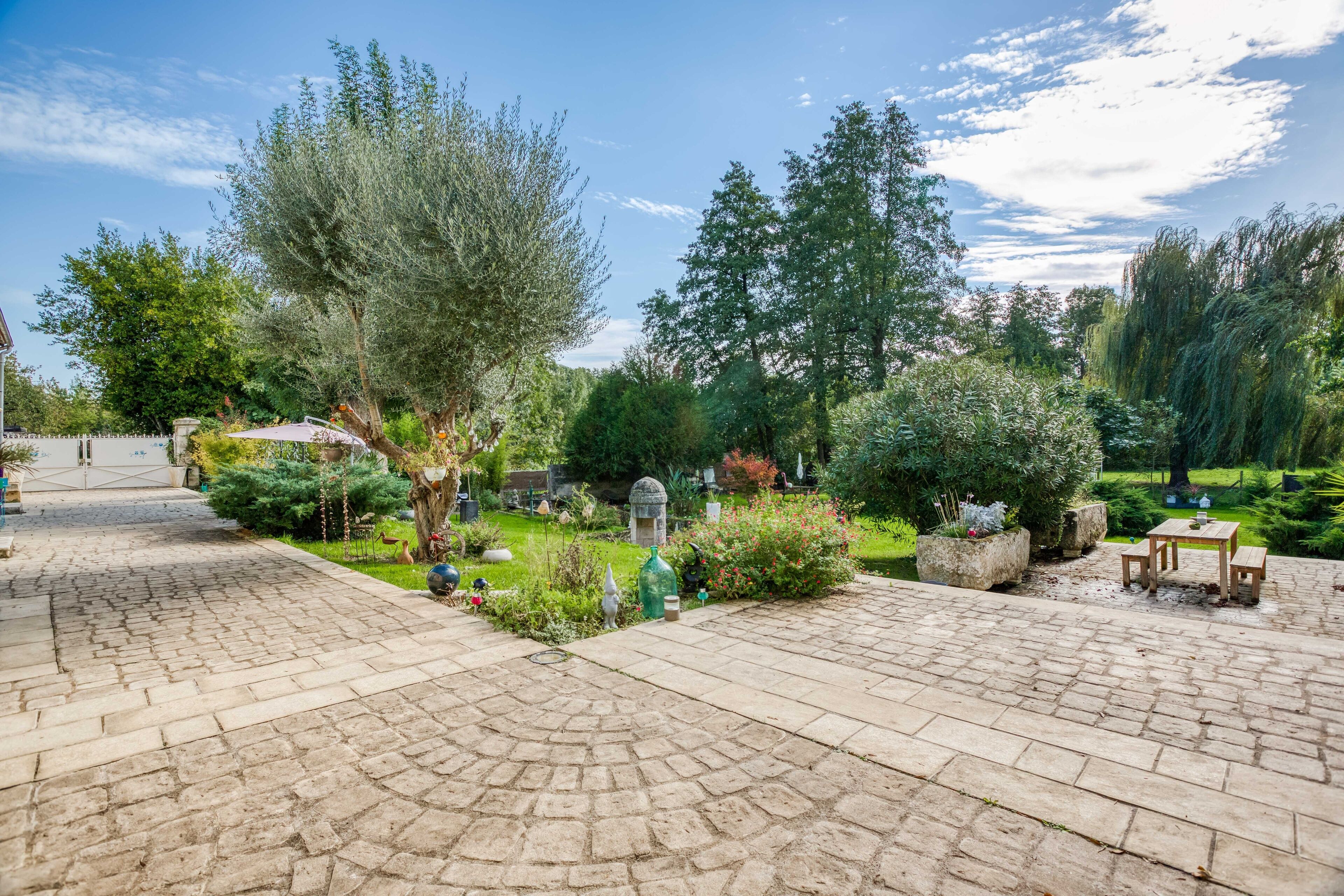 Une cour dallée, recouverte de pierres, entourée d’une végétation luxuriante et d’arbres. On y trouve une petite table et un banc au centre.