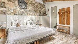 A bedroom with a stone wall, a bed with patterned bedding, a wooden bench, and a coat rack. - Jolysable (Pons)
