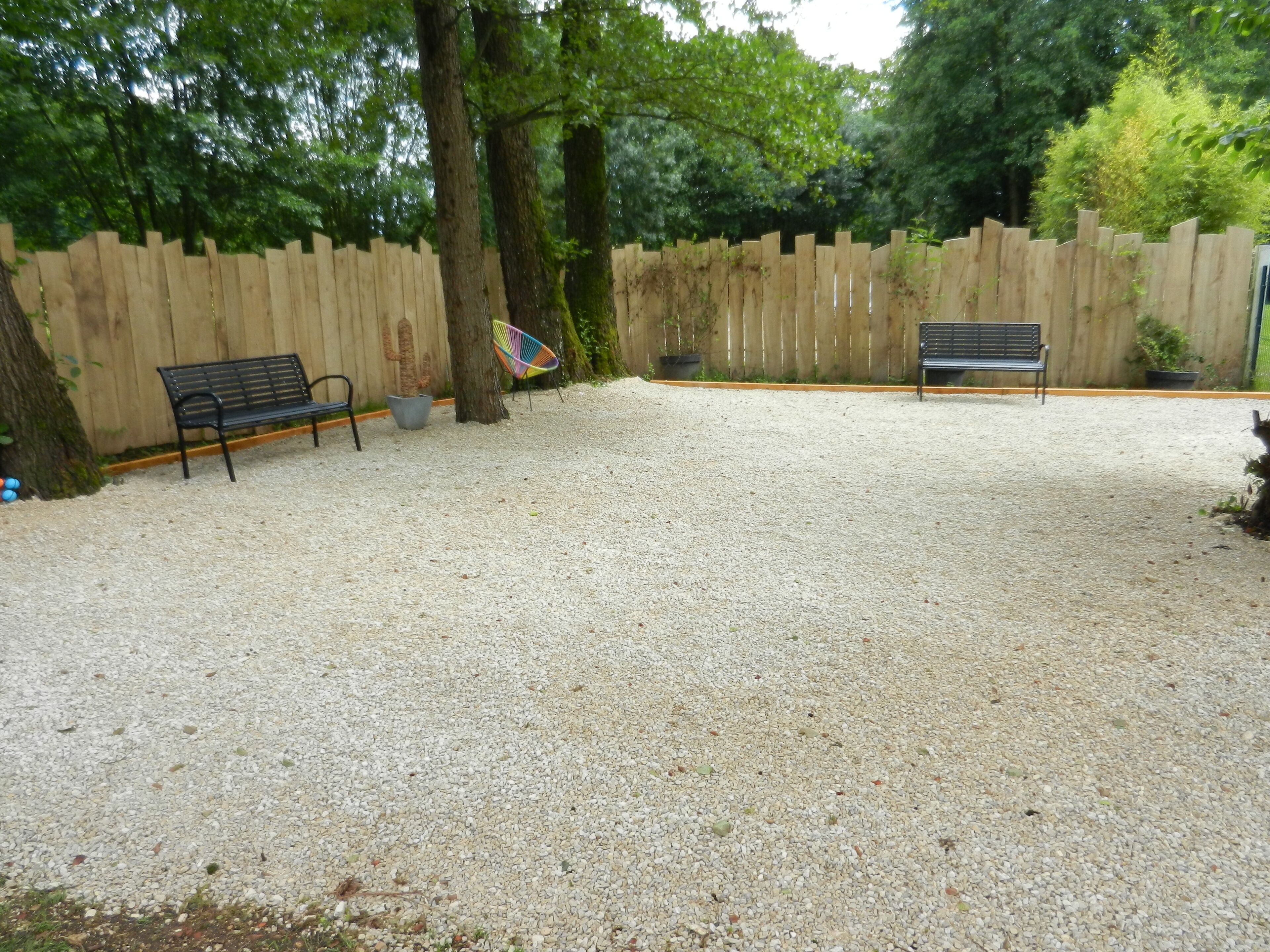 Een buitengebied met grind, twee banken, een houten omheining en bomen.