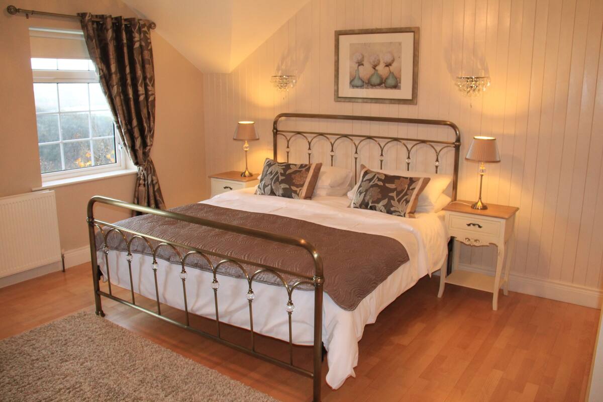 A bedroom with a metal bed frame, white bedding, two bedside tables with lamps, a window with curtains, and a framed picture on the wall.